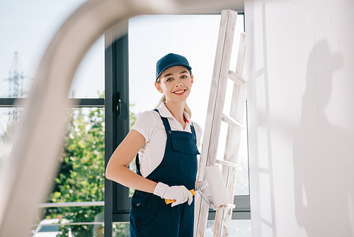 selective focus of pretty young painter in uniform holding paint roller and smiling at camera