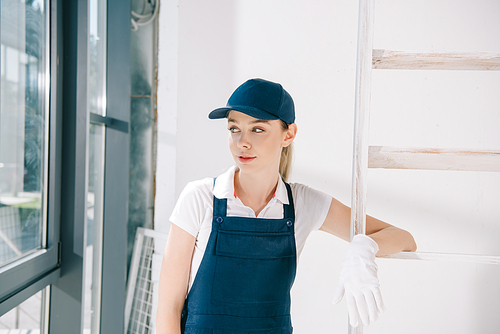 attractive young painter in uniform standing near ladder and looking away