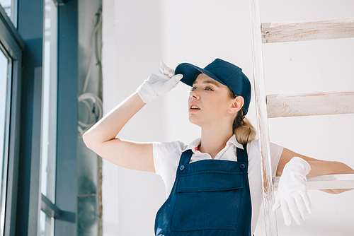 pretty young painter in uniform touching cap while looking away