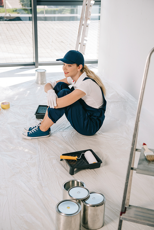 beautiful young painter in overalls and cap sitting on floor near paint roller in roller tray and cans with paint