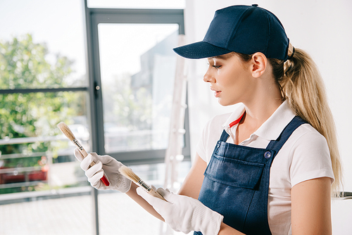 beautiful young painter in overalls and cap holding paintbrushes