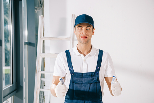 handsome painter in uniform showing thumbs up and smiling at camera