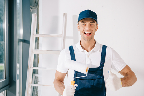 handsome young painter in uniform holding paint roller, showing thumb up and smiling at camera