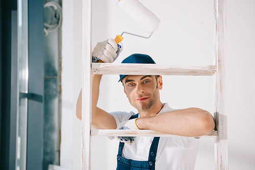 handsome young painter in uniform standing near ladder, holding paint roller and smiling at camera