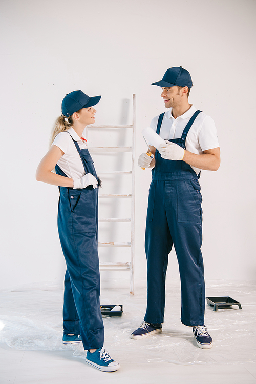 handsome painter in uniform holding paint roller while talking to pretty colleague