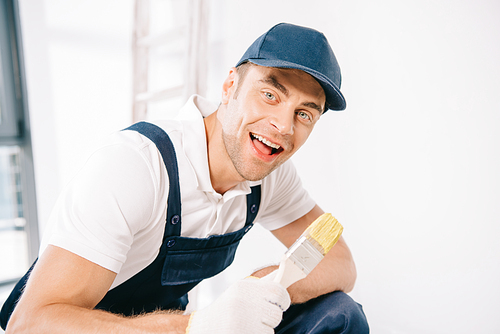 cheerful young painter in uniform holding paintbrush and smiling at camera