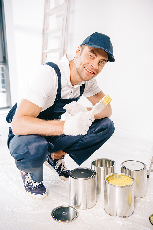 handsome young painter in uniform holding paintbrush near cans with paint and smiling at camera