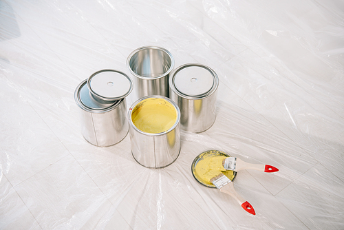 cans with paint and yellow paintbrushes on white floor covered with cellophane