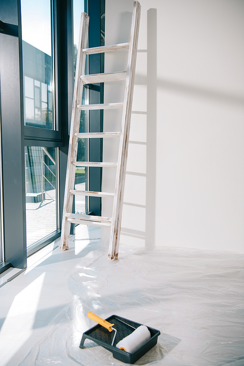 room with large window, ladder near white wall, paint roller in roller tray on floor covered with cellophane