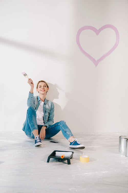 happy young woman sitting under white wall with painted pink near and smiling at camera