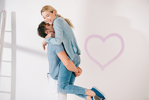 happy young man holding girlfriend on hands near pink heart drawn on white wall