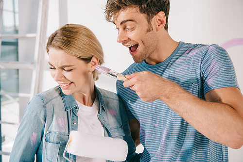 cheerful young man having fun with girlfriend while frightening with pink paintbrush