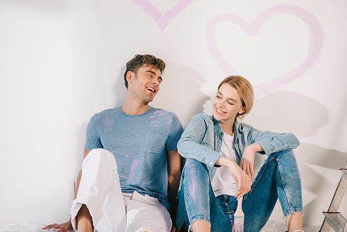 happy young couple sitting by white wall under drawn pink heart