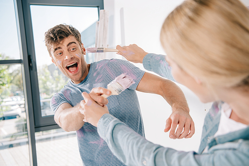 cheerful young couple having fun and fighting on pink paintbrushes while making room repair