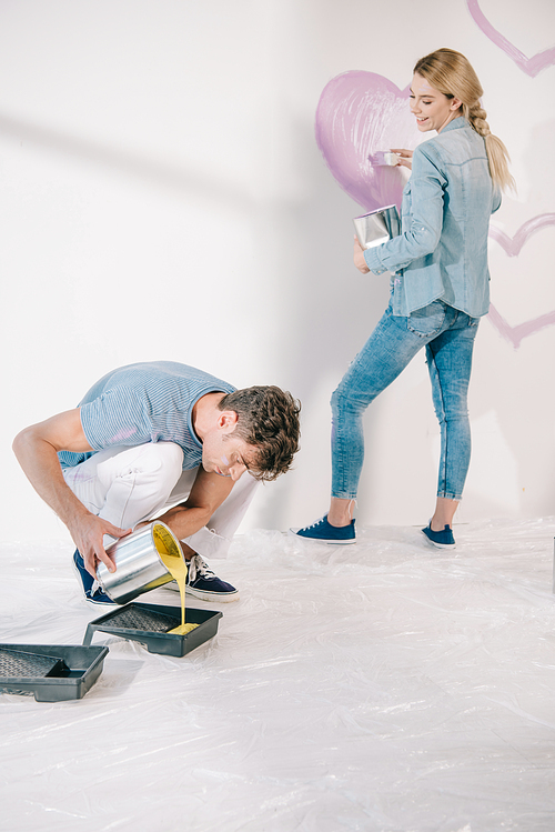 young man adding yellow paint into roller tray while girlfriend drawing pink heart on white wall