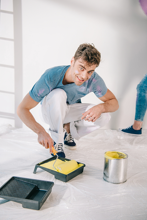 handsome young man putting paint roller into roller tray with yellow paint and smiling at camera