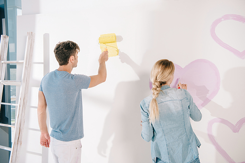 man painting white wall in yellow while girlfriend drawing pink heart