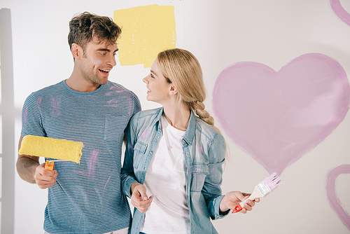 happy young couple looking at each other while standing near pink heart painted on white wall