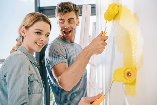 cheerful young couple painting wall in yellow with paint rollers