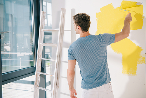 back view of young man painting white wall in yellow with paint roller