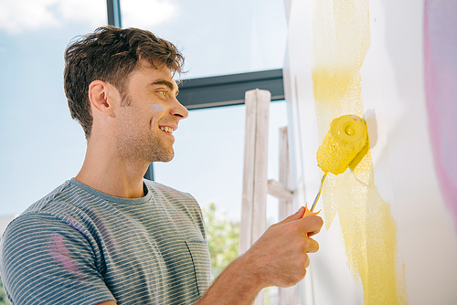 handsome young man smiling while painting wall in yellow with paint roller