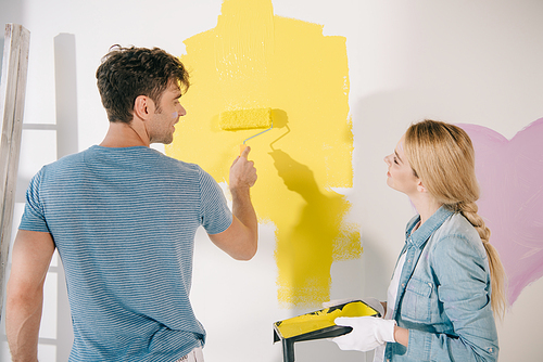 young woman holding roller tray while boyfriend painting wall in yellow with paint roller