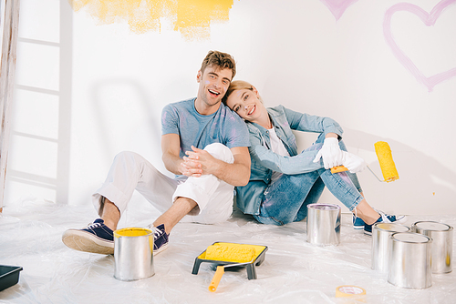 happy young woman holding yellow paint roller while sitting on floor near boyfriend