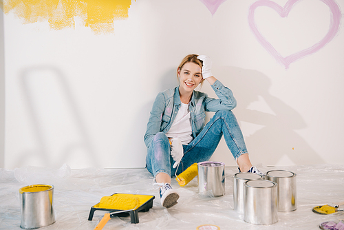 attractive young woman holding yellow paint roller while sitting on floor near wall