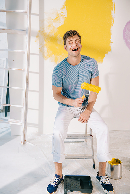 handsome young man sitting on ladder, holding yellow paint roller and smiling at camera