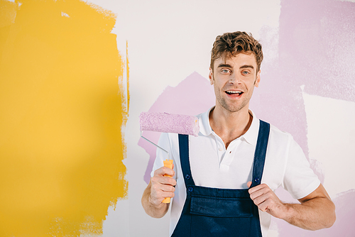 handsome young painter in overalls holding paint roller while standing near wall painted in yellow and pink