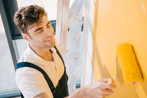 handsome young painter smiling while painting wall in yellow color with paint roller