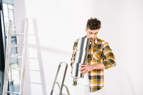 handsome young man holding cans with paint while standing near white wall