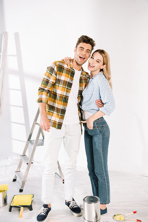 happy young couple embracing and smiling at camera while standing near white wall