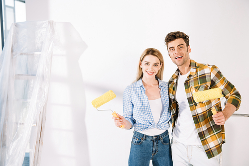 happy young couple smiling at camera while holding yellow paint rollers