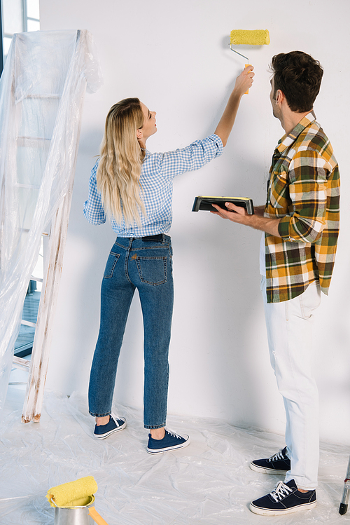 young woman painting wall in yellow while boyfriend holding roller tray