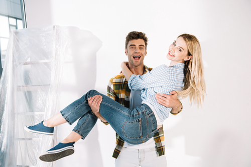 happy young man holding girlfriend on hands while standing near white wall