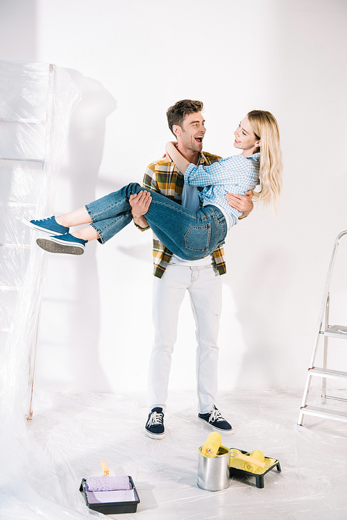 happy young man holding girlfriend on hands while standing near white wall