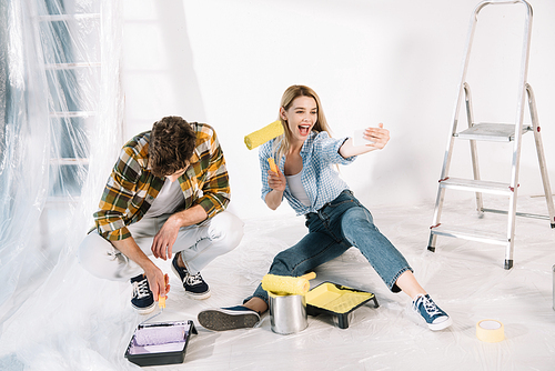 cheerful young woman holding yellow paint roller and taking selfie while boyfriend putting paint roller into pink paint