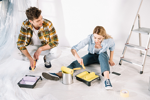 handsome man holding pink paint roller and woman putting paint roller into yellow paint while sitting on floor