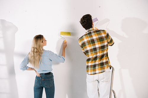 young woman holding yellow paint roller and boyfriend holding pink paint roller white standing near white wall