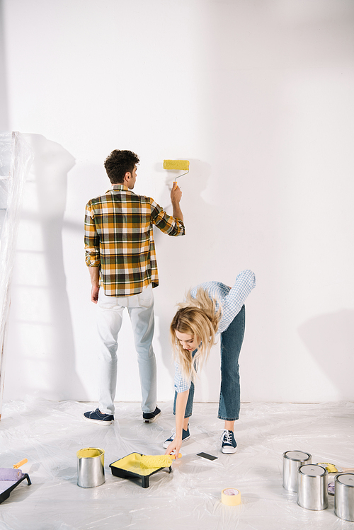 young woman putting paint roller into roller tray with yellow paint while boyfriend painting wall