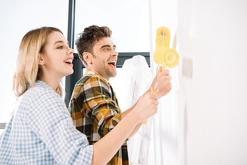 happy young couple painting white wall in yellow with paint rollers