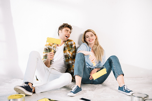 happy young couple sitting near white wall and holding yellow paint rollers