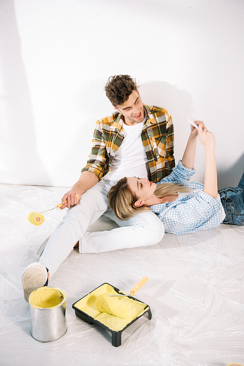 young woman taking selfie with boyfriend holding yellow paint roller while sitting on floor