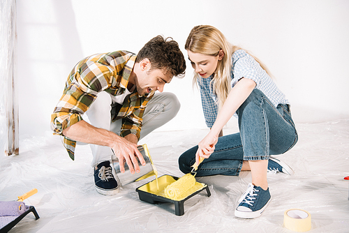 young man adding yellow paint into roller tray while girlfriend holding paint roller