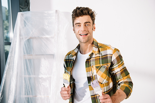 cheerful young man  while holding yellow and pink paintbrushes