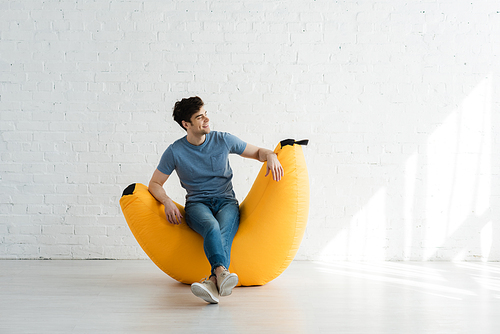 happy man sitting with crossed legs on yellow bean bag chair near white brick wall