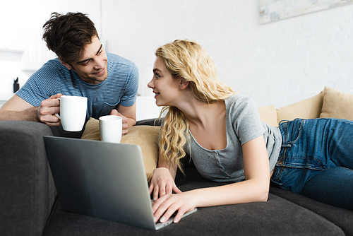 happy and handsome man holding cups near blonde girl using laptop