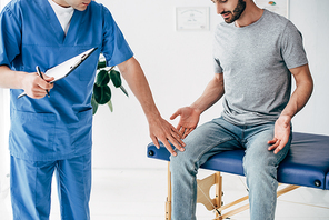 cropped view of doctor examining patient knee and holding clipboard in massage cabinet at clinic