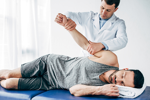 chiropractor stretching arm of handsome patient lying on massage table in hospital
